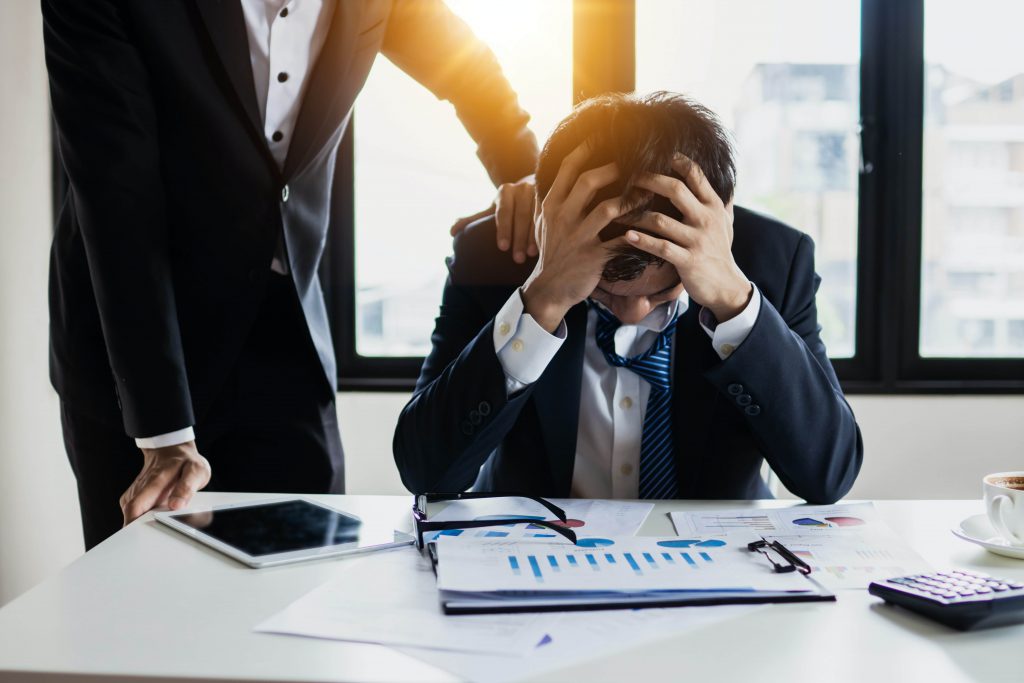 A stressed out employee with his heads in his hands and a coworker with a shoulder on his arm